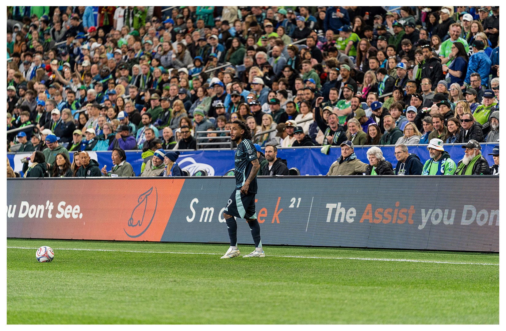 A Seattle Sounders player on the pitch, with SmarTek21 signage and supporters behind him