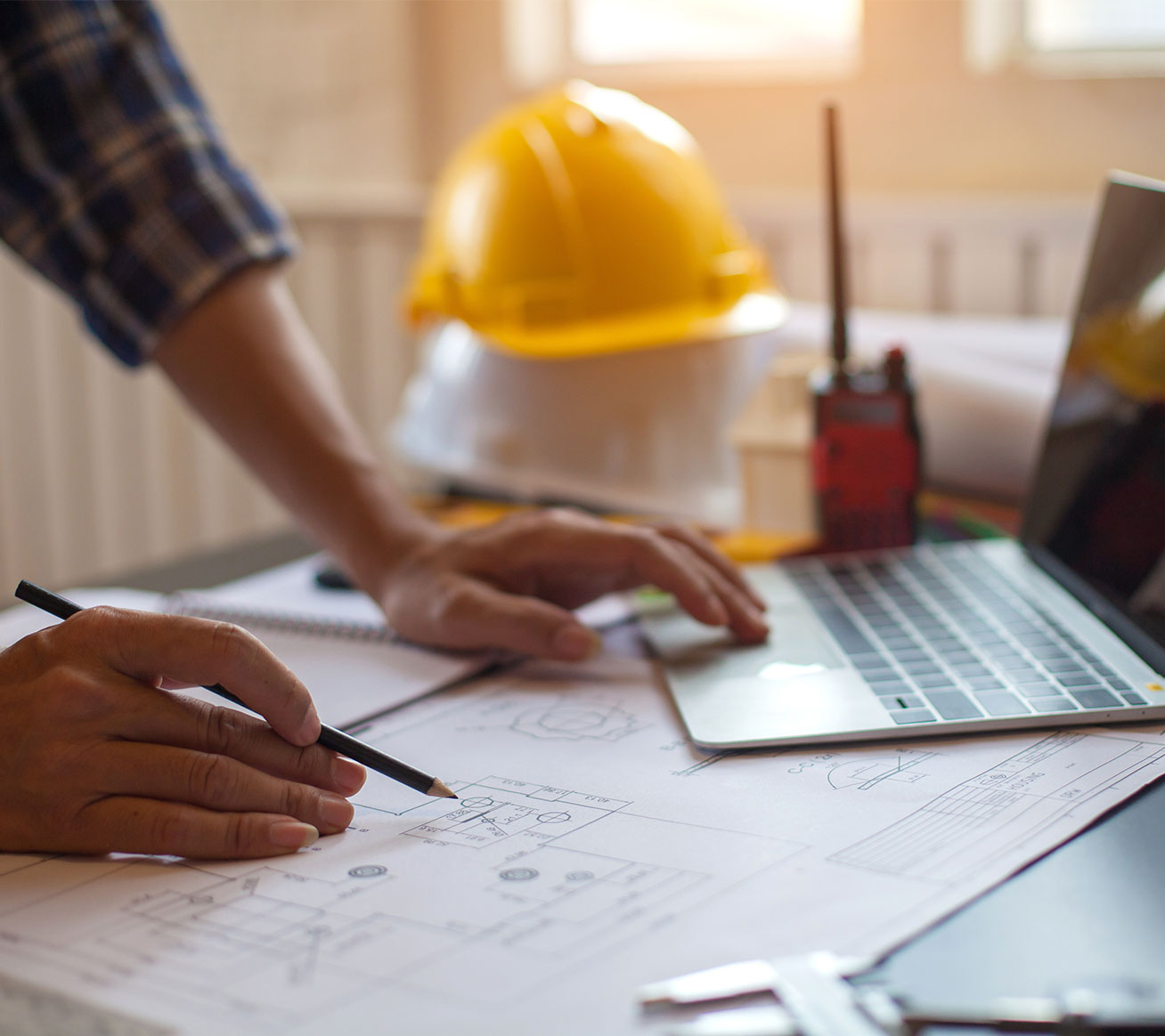 Hands working on building plans beside a laptop and yellow hard hat