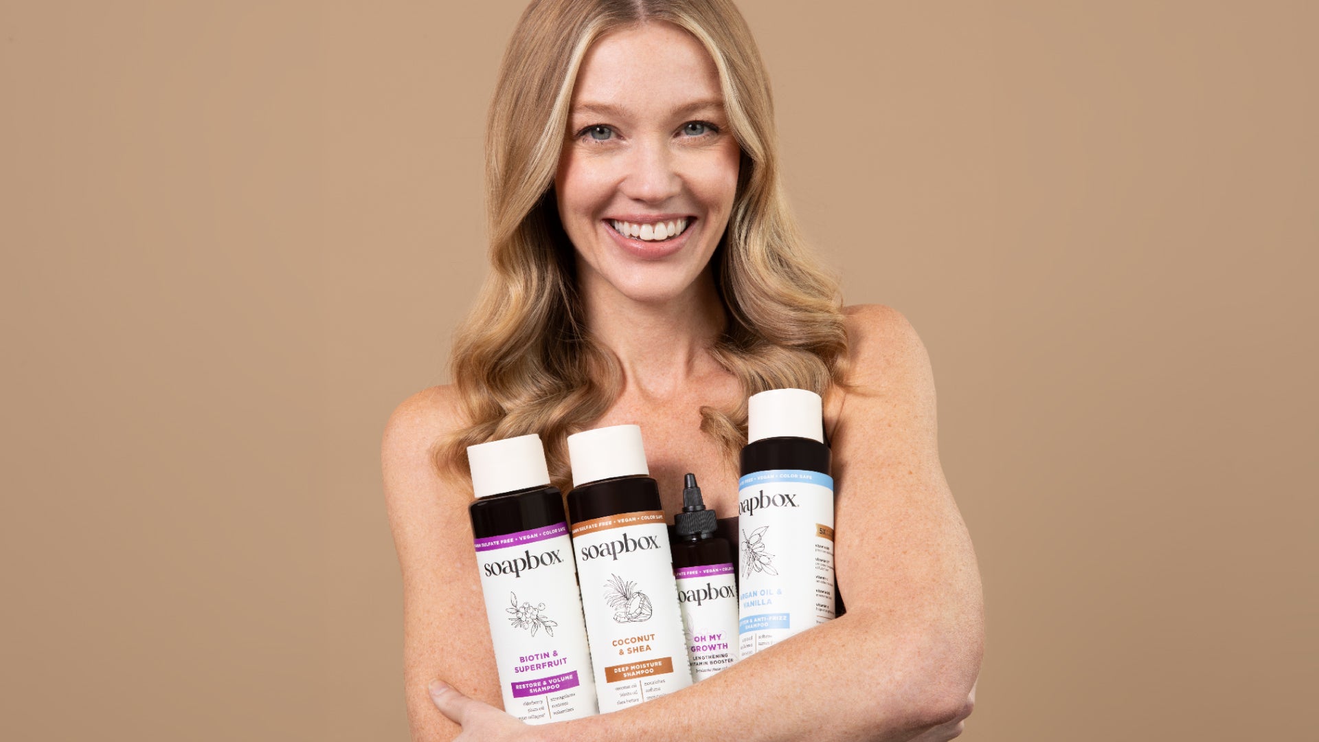 A woman holding four Soapbox hair-care products against a warm brown background