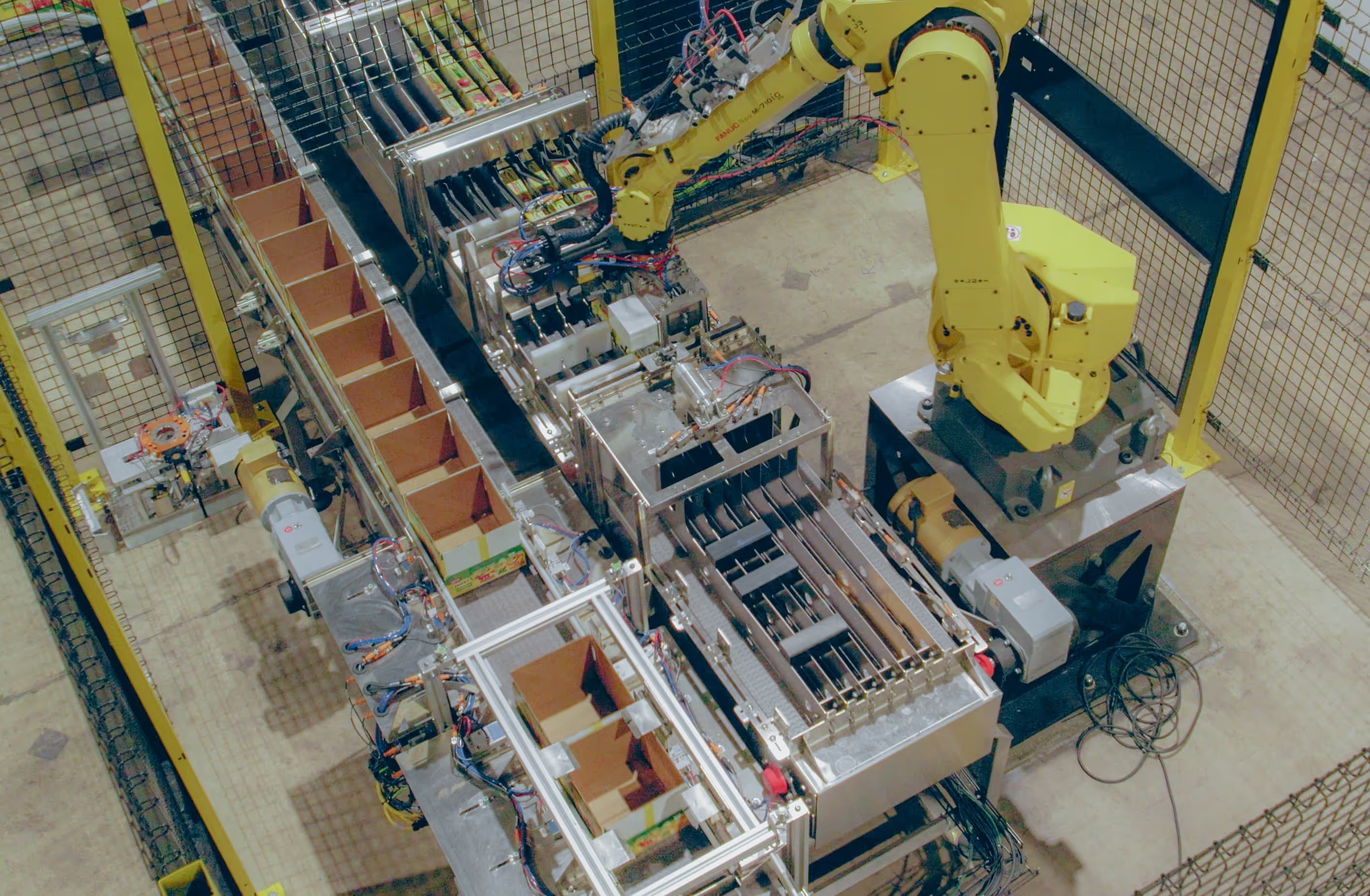 An overhead view of a yellow industrial robot beside conveyors and open cardboard cases at Sojo