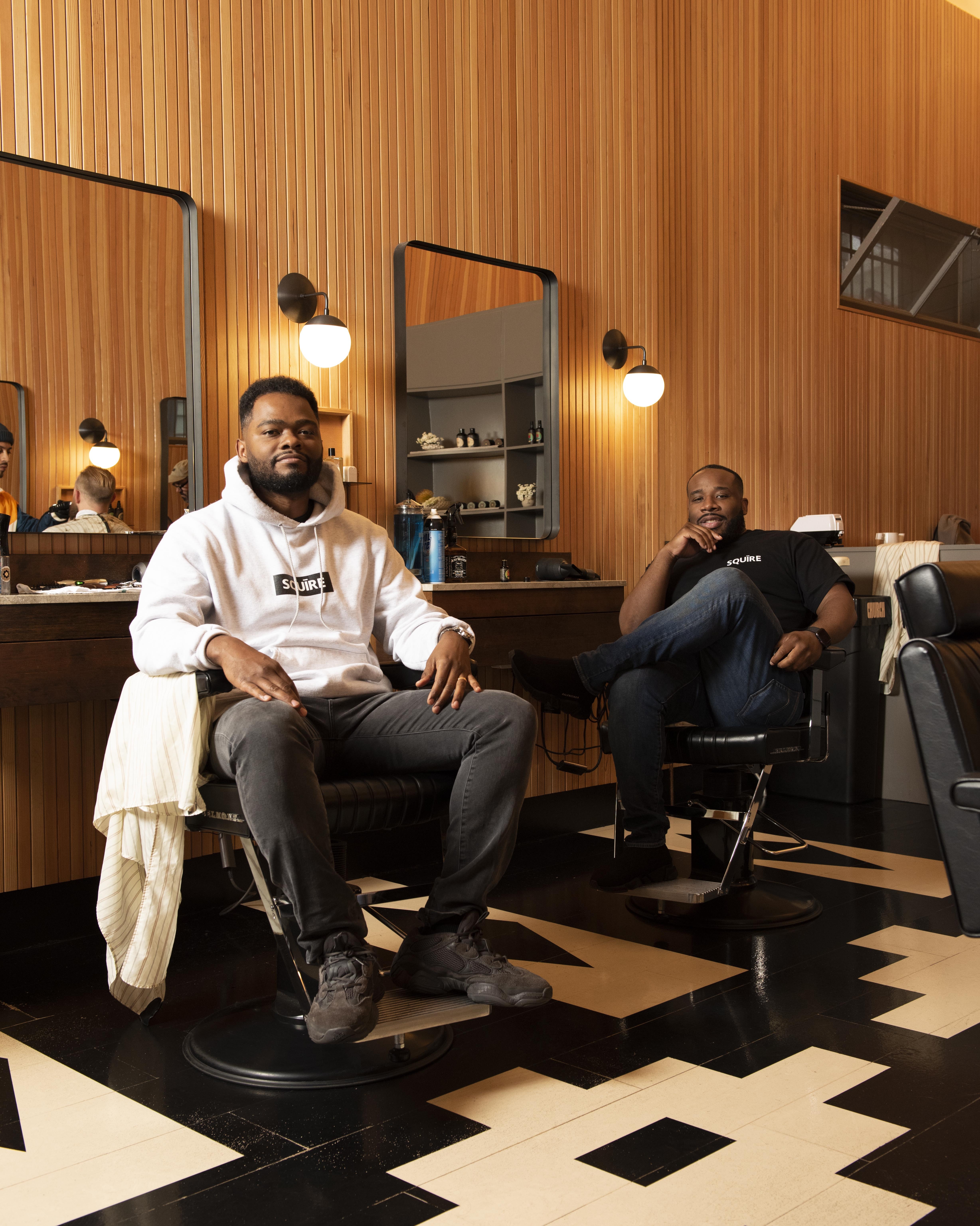 Songe LaRon and Dave Salvant seated in barber chairs inside a wood-paneled barbershop