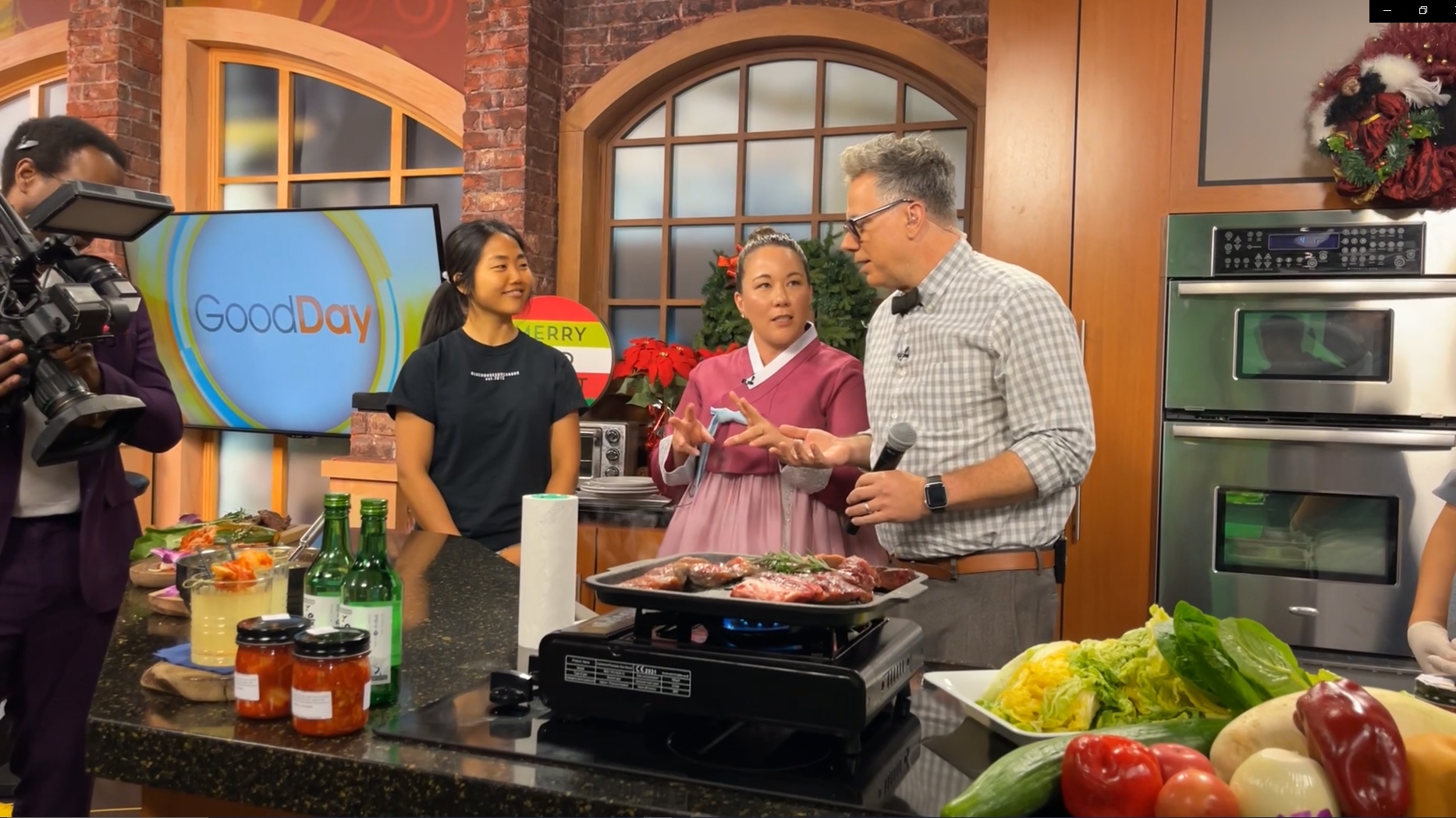 Sonya Harris, wearing a pink hanbok, speaking with a presenter beside a Korean food demonstration on the GoodDay television set.