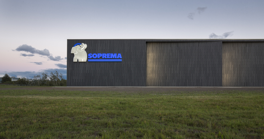 A dark SOPREMA manufacturing building set behind a grassy field in Canada