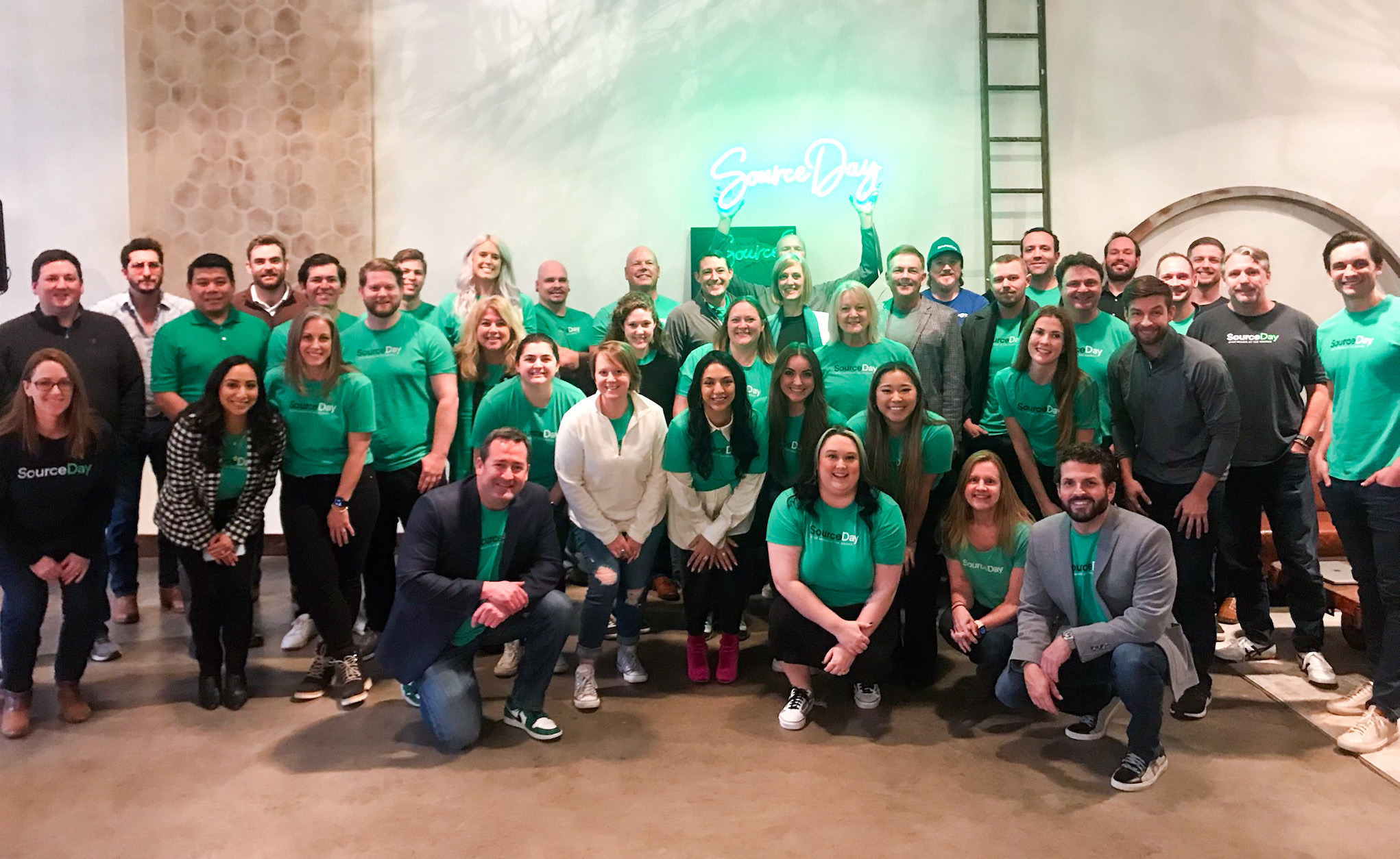 SourceDay employees gathered in green company shirts beneath a neon SourceDay sign