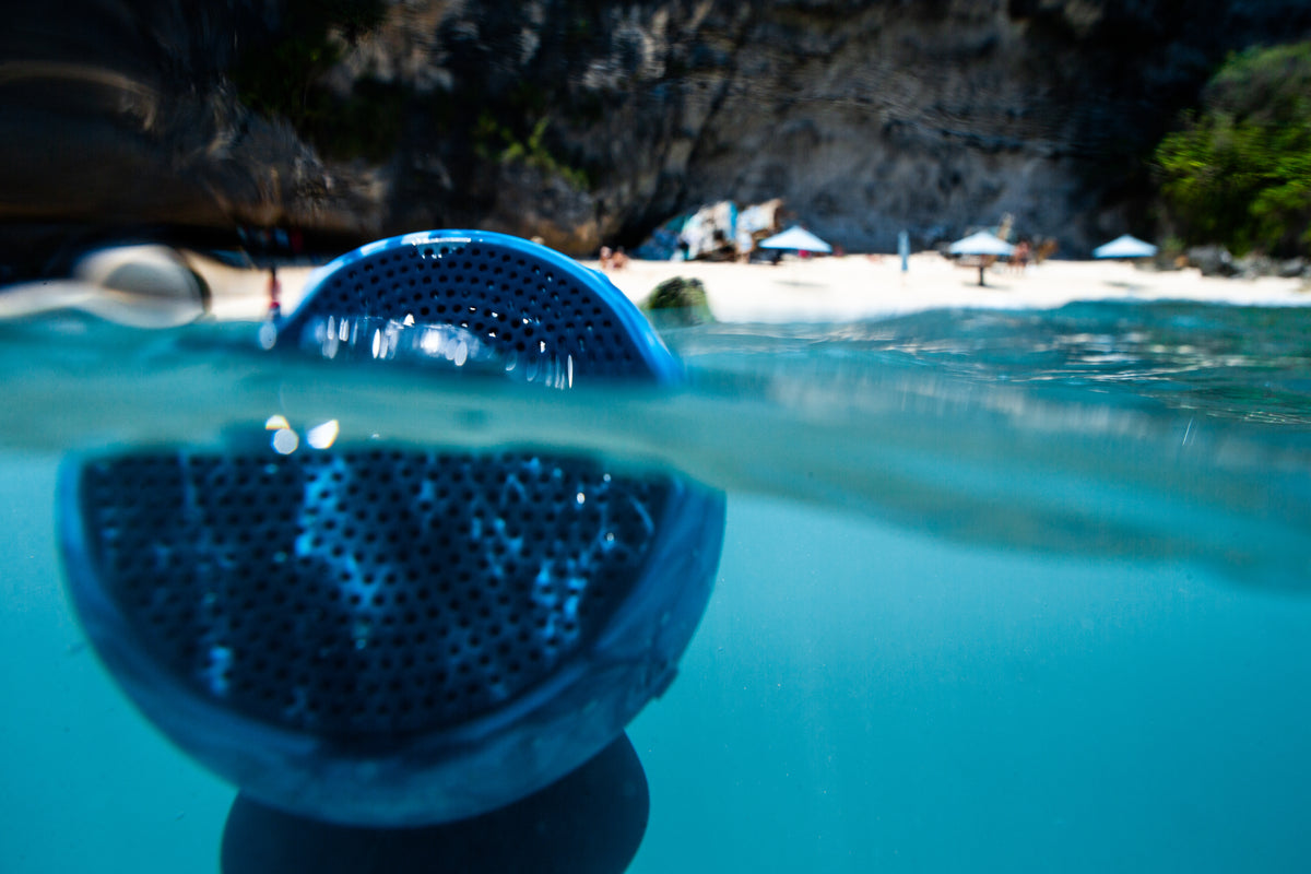 A Speaqua Barnacle speaker floating half-submerged in clear water off a beach