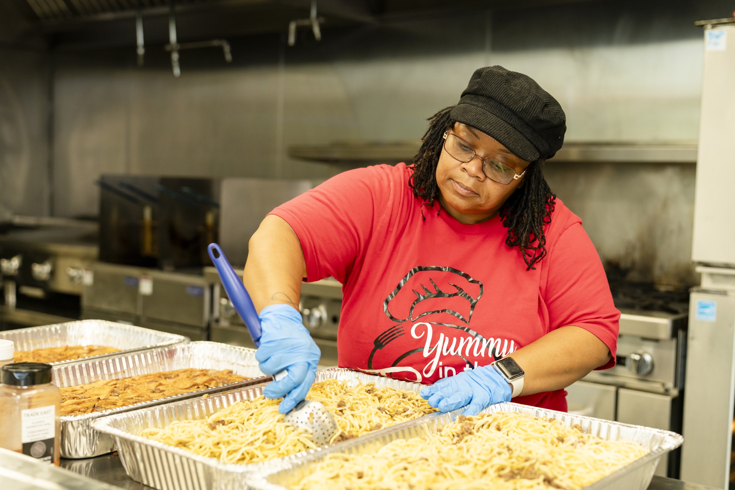 A food entrepreneur wearing blue gloves prepares large trays of food in a commercial kitchen