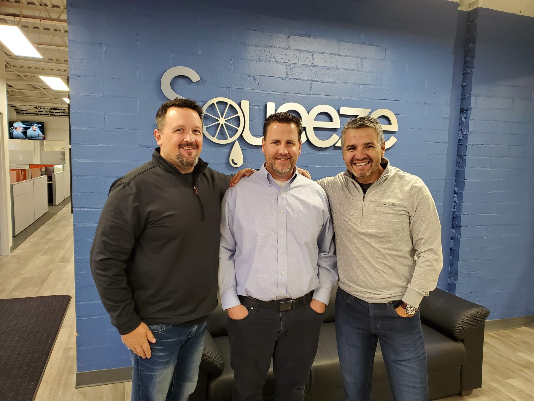 Three people pose in a Squeeze office beside its citrus-themed wall sign