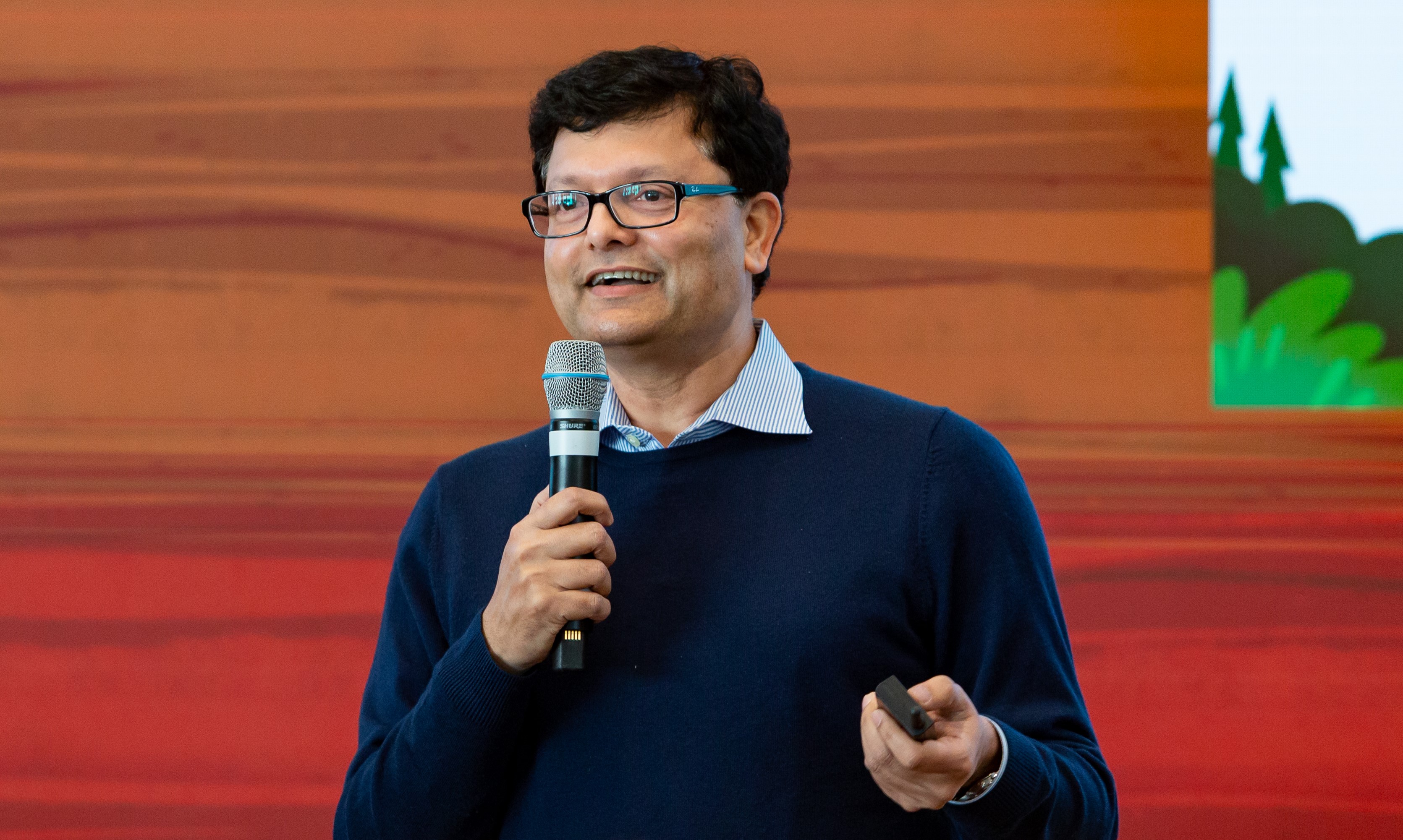 Srini Tallapragada speaking on stage, holding a microphone and presentation remote
