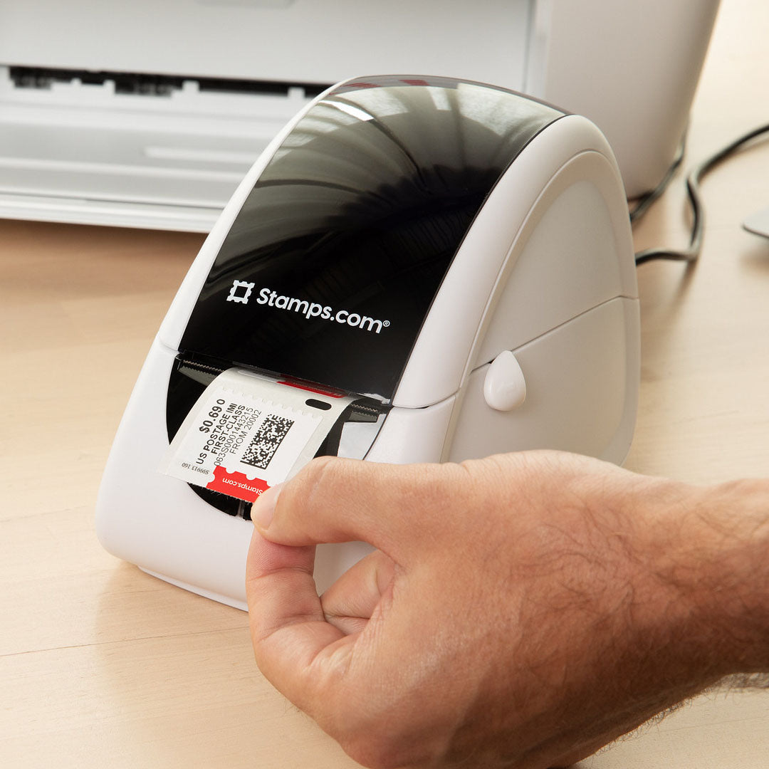 A hand removes printed postage from a Stamps.com thermal printer on an office desk