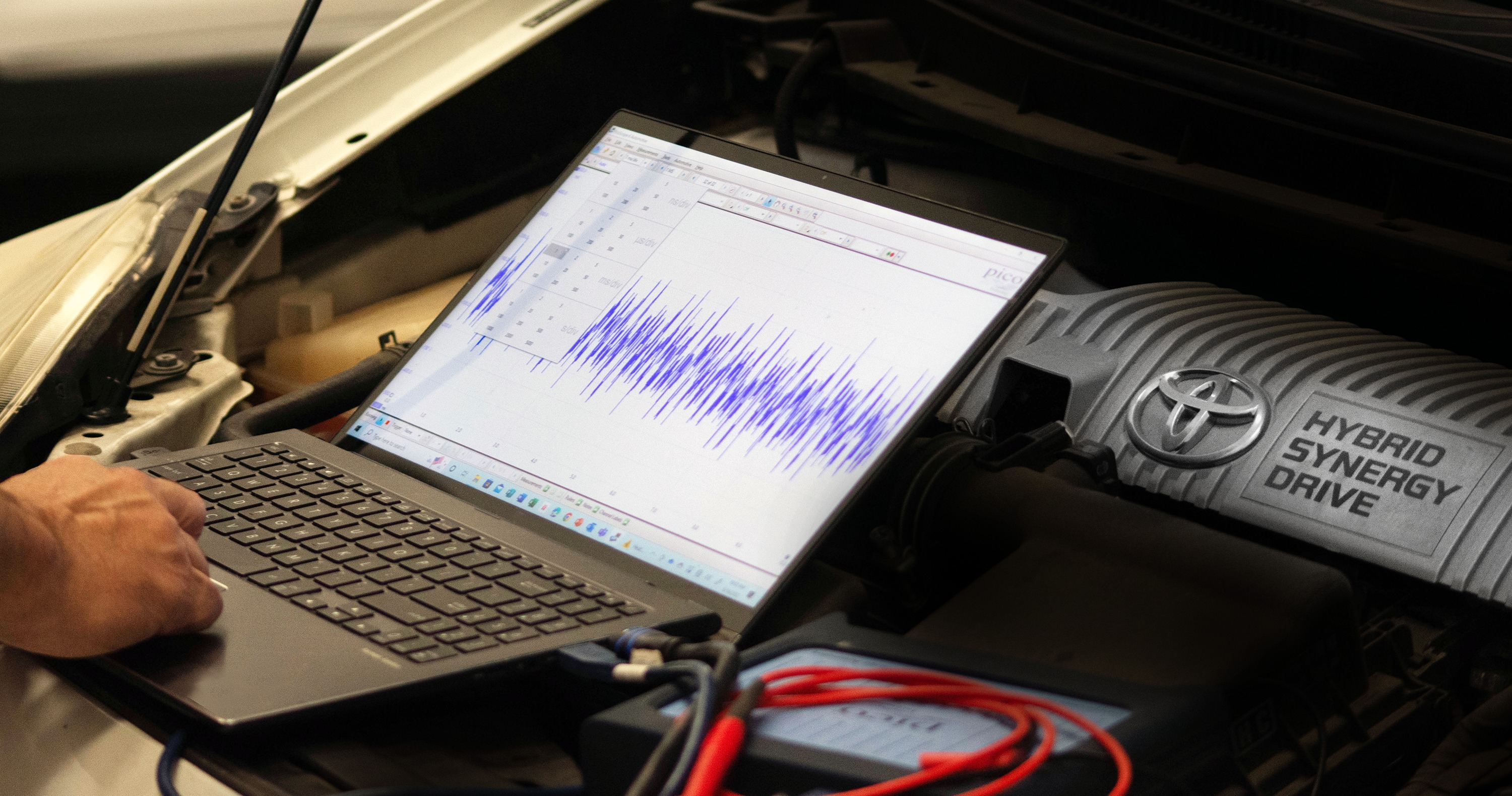A laptop displays diagnostic waveforms beside a Toyota hybrid engine