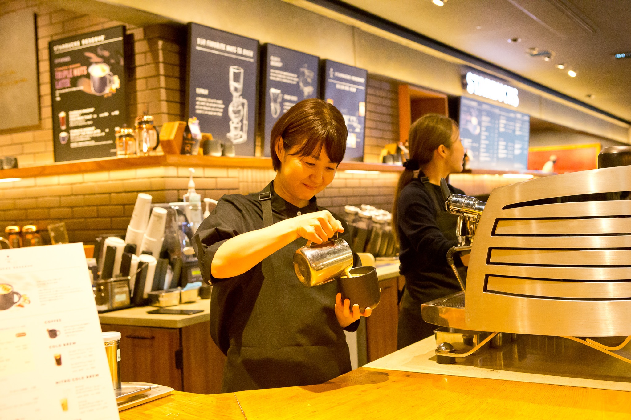 A Starbucks Japan barista pours steamed milk into a ceramic cup while a colleague works beside the espresso machine