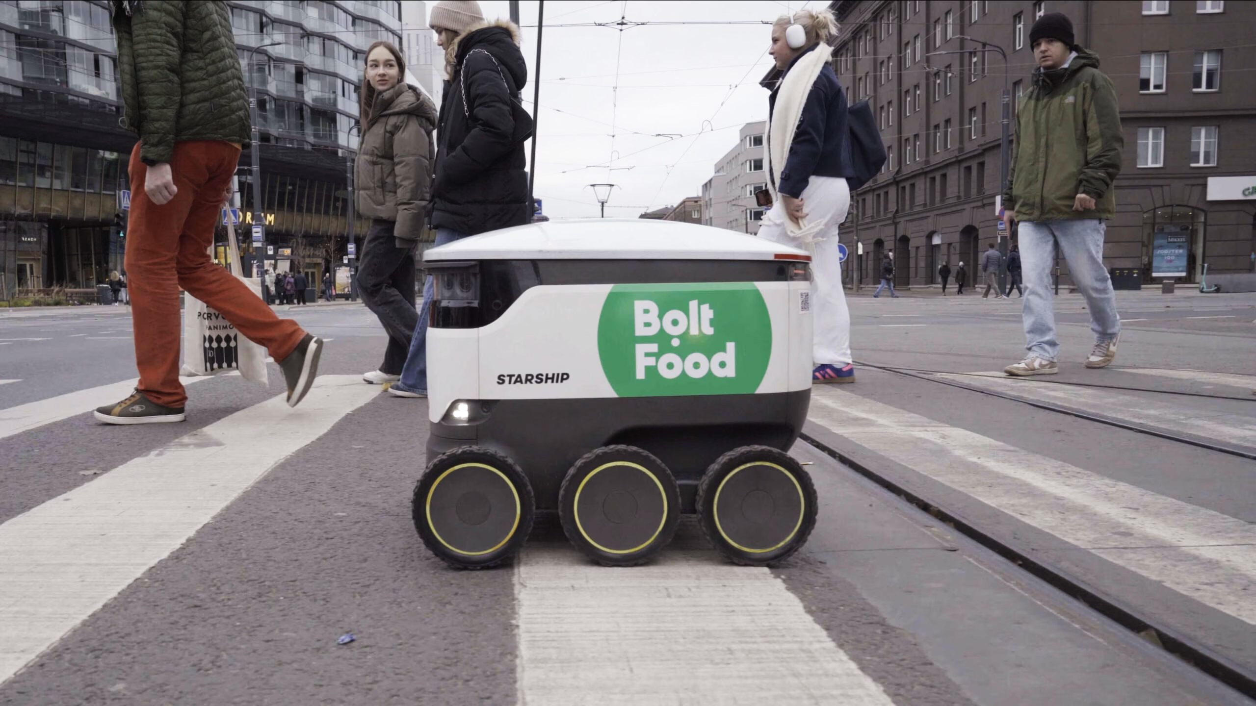 A Bolt Food branded Starship robot waits at a city crossing among pedestrians