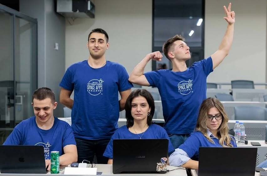 Five hackathon participants in blue NASA Space Apps shirts, three working at laptops and two standing behind them