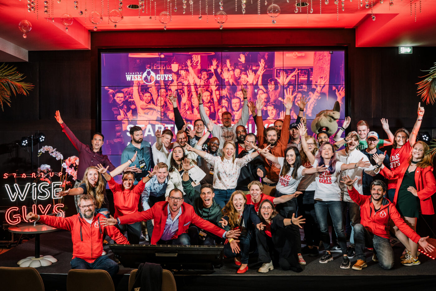 Startup Wise Guys community gathered on a stage, many wearing red and raising their arms