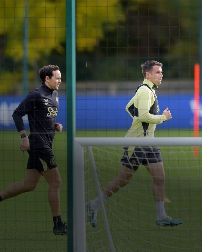 Two people running on a training pitch, with the foreground player wearing a tracking vest