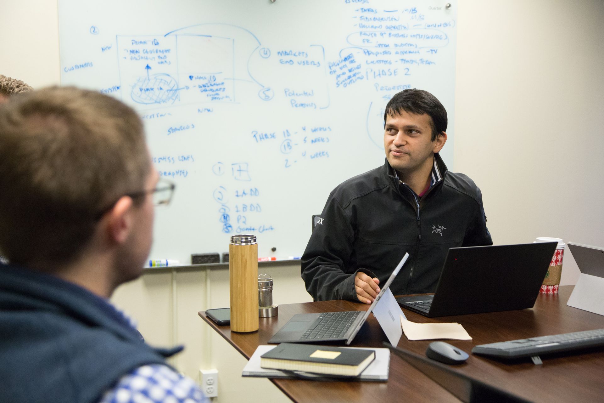 Stax colleagues discussing work around a table with laptops and a marked-up whiteboard