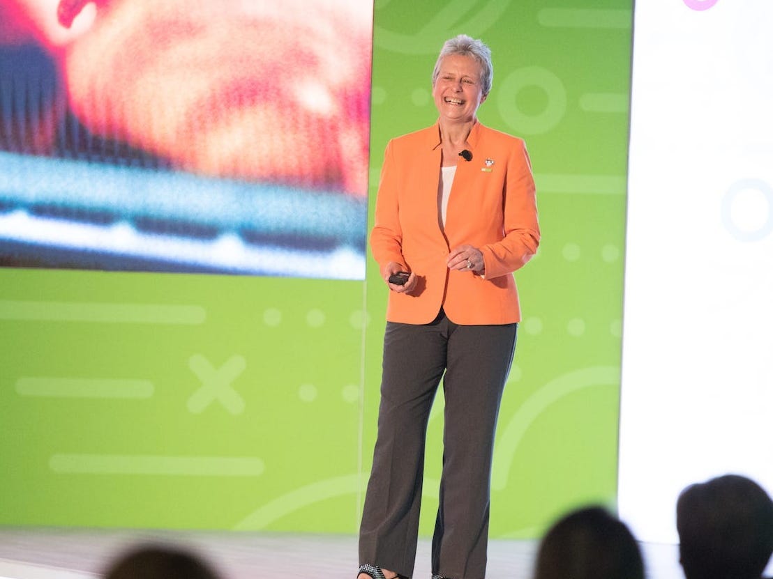 Stefania Mallett speaking onstage in an orange jacket, holding a presentation clicker