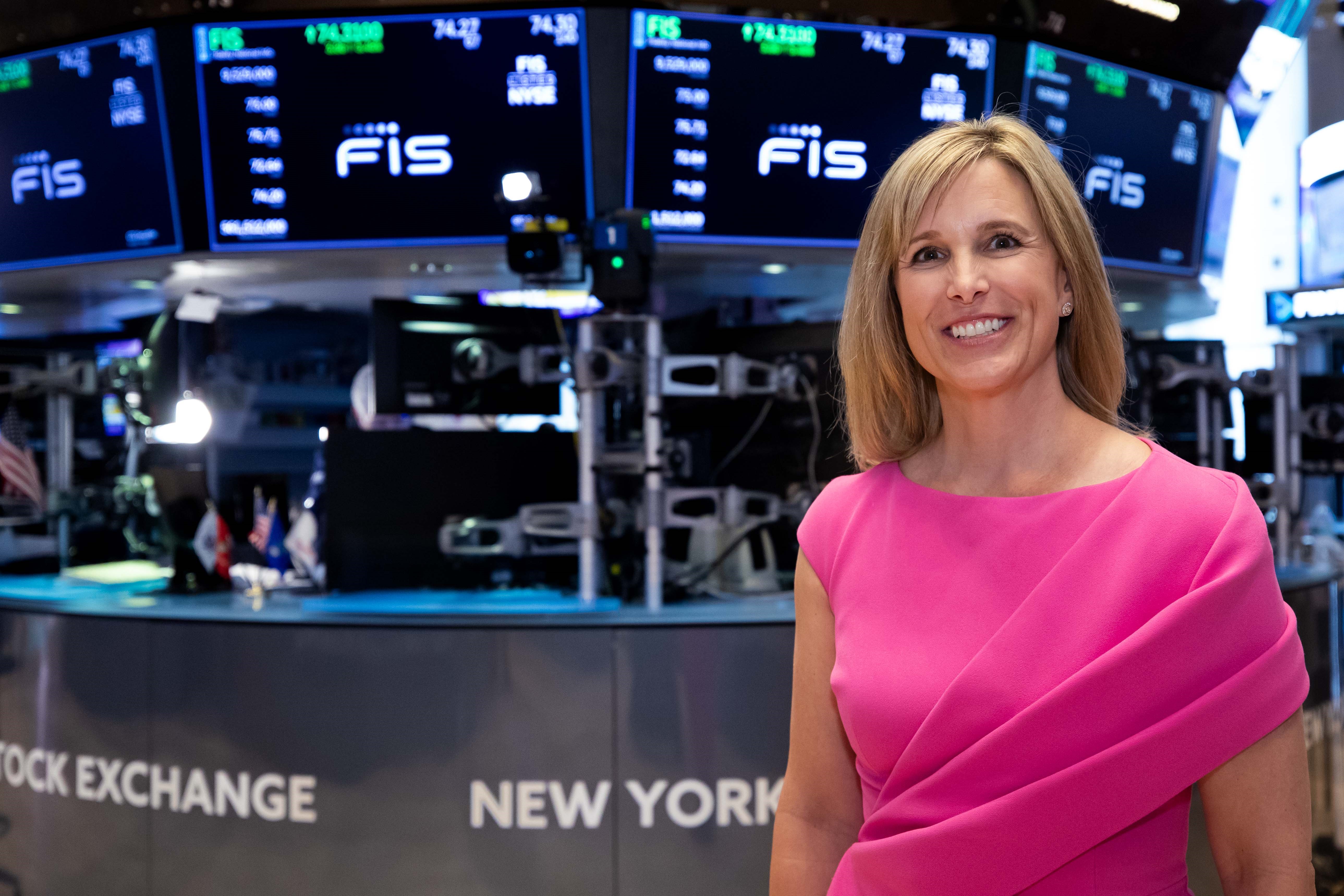 Stephanie Ferris in a pink dress on the New York Stock Exchange trading floor, with FIS screens behind her