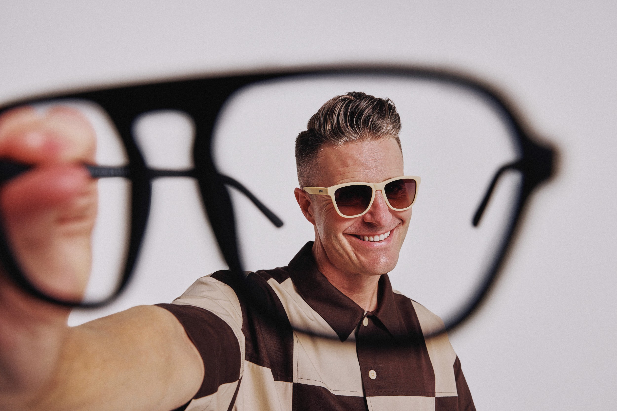 Stephen Lease holds an oversized black sunglasses frame toward the camera while wearing a cream pair