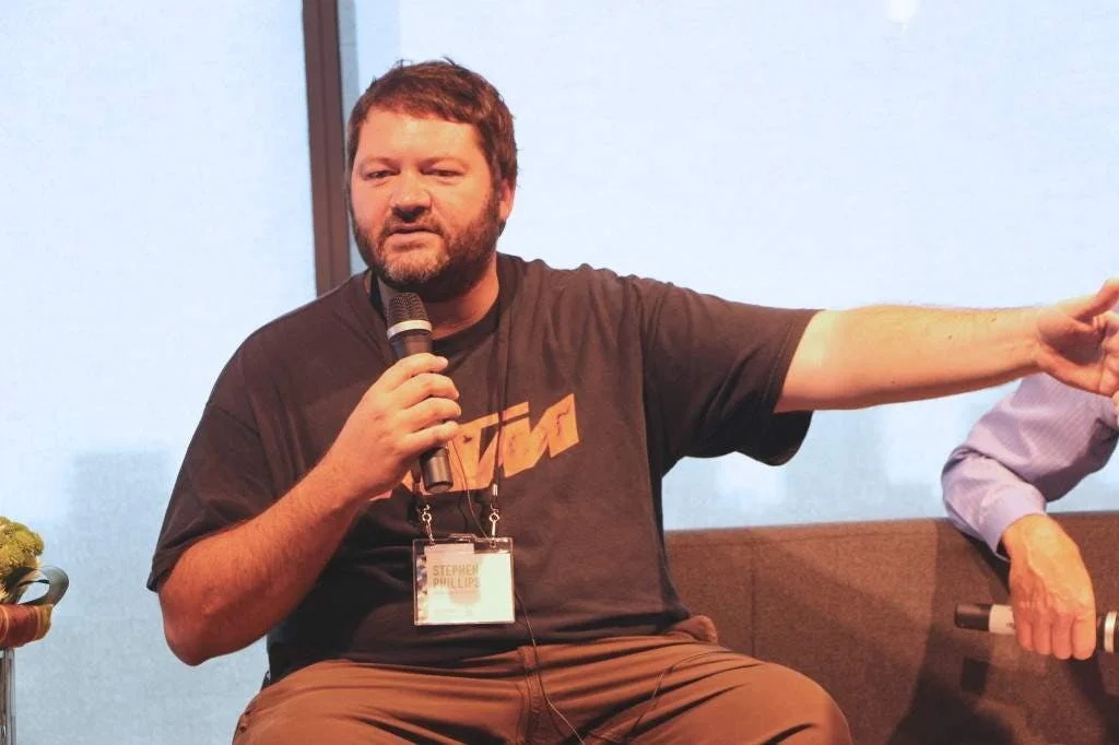 Stephen Phillips seated with a microphone, speaking and gesturing during a public appearance