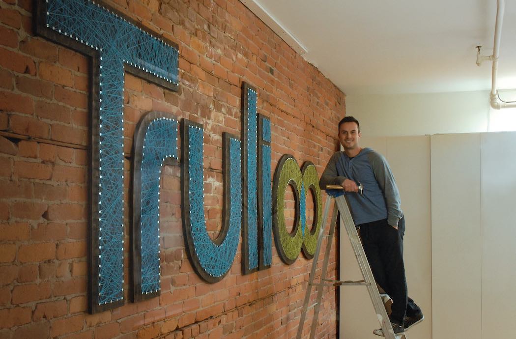 Stephen Ufford beside a large Trulioo sign, leaning against a ladder in the company’s office