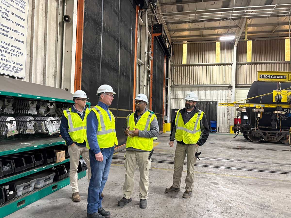 Steve Angel wearing a hard hat and yellow safety vest, talking with CSX employees in a railway workshop.