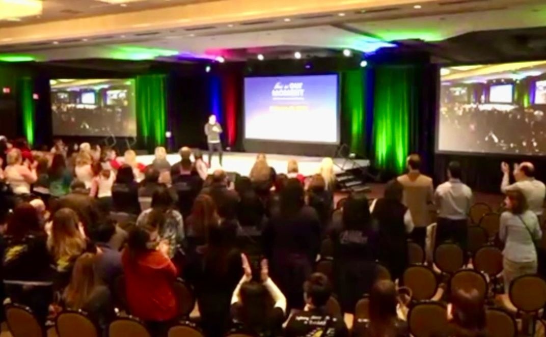 Audience standing in a conference room during Steve Bilt's first meeting after his return