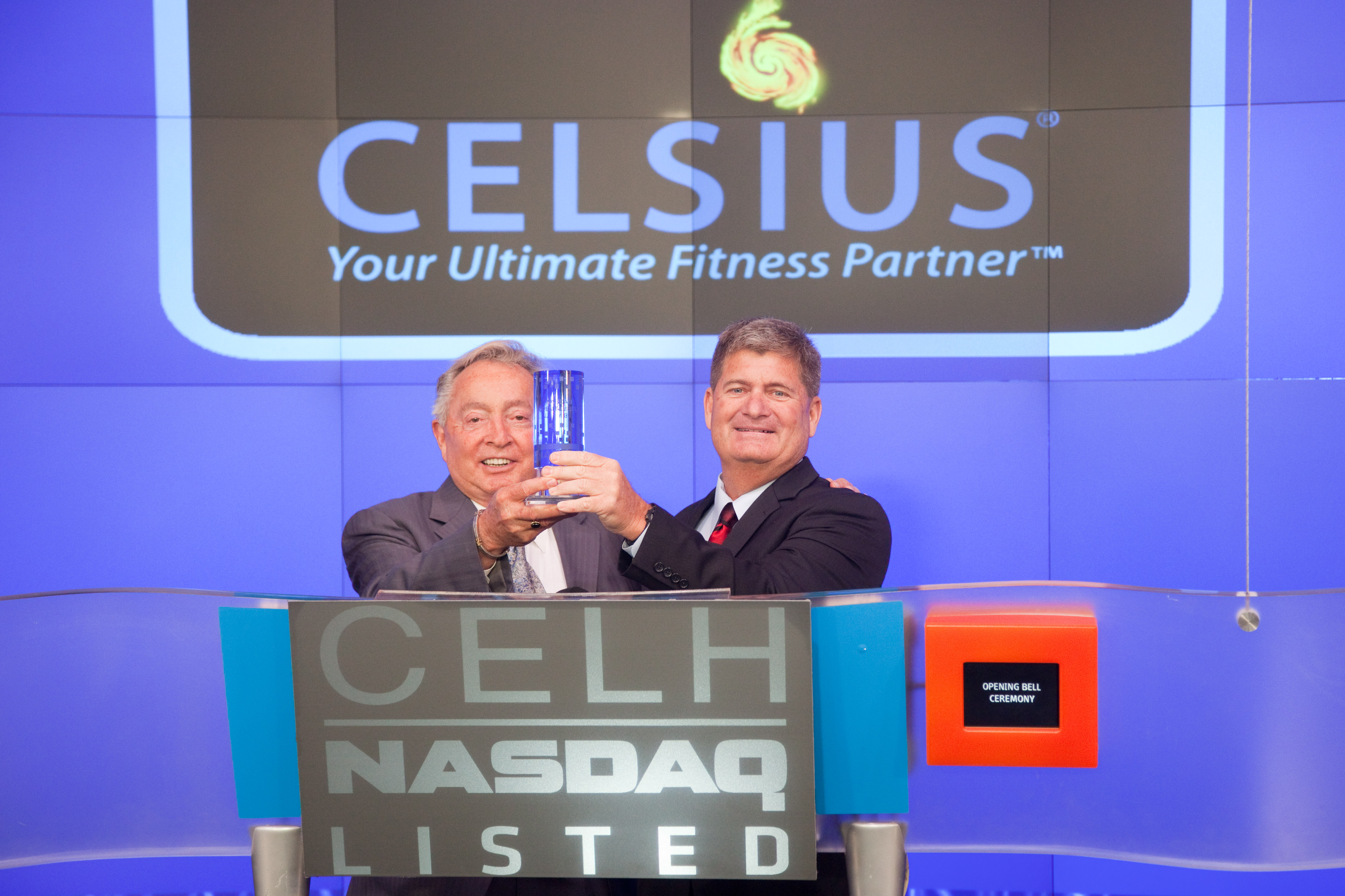 Carl DeSantis and Steve Haley holding a trophy at the Nasdaq opening-bell podium