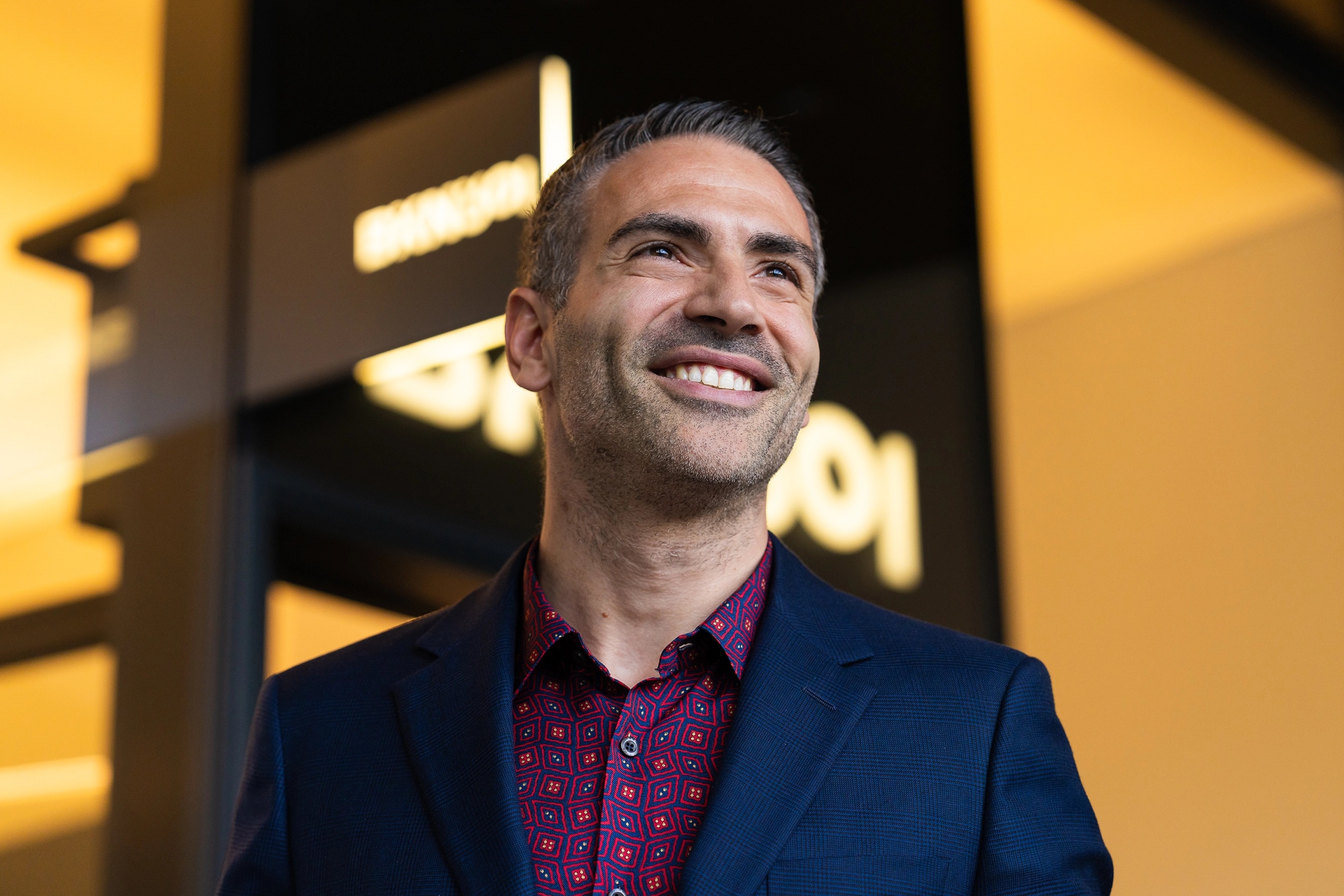 Stiven Muccioli smiling in a blue jacket and patterned shirt in front of BKN301 signage
