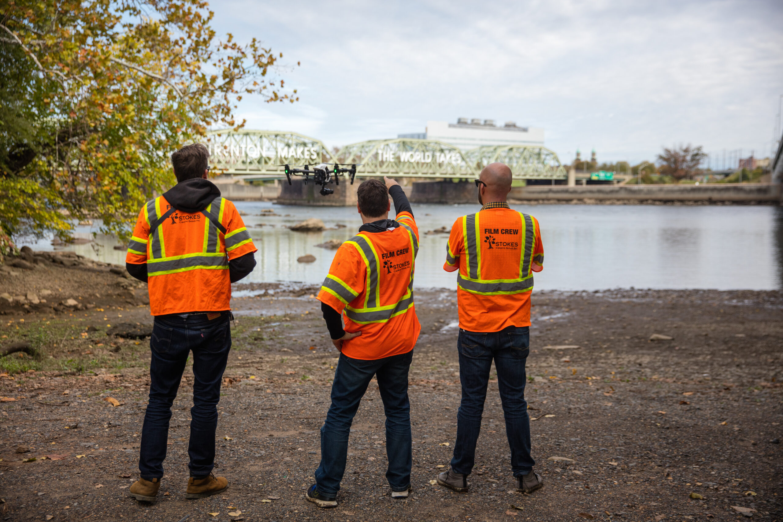 Three Stokes Creative Group crew members in safety vests operate a drone beside a river and bridge