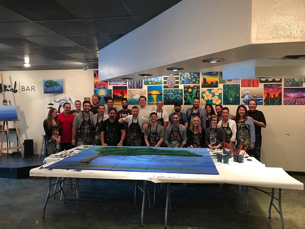 A group in aprons gathers behind a shared painting at an art studio