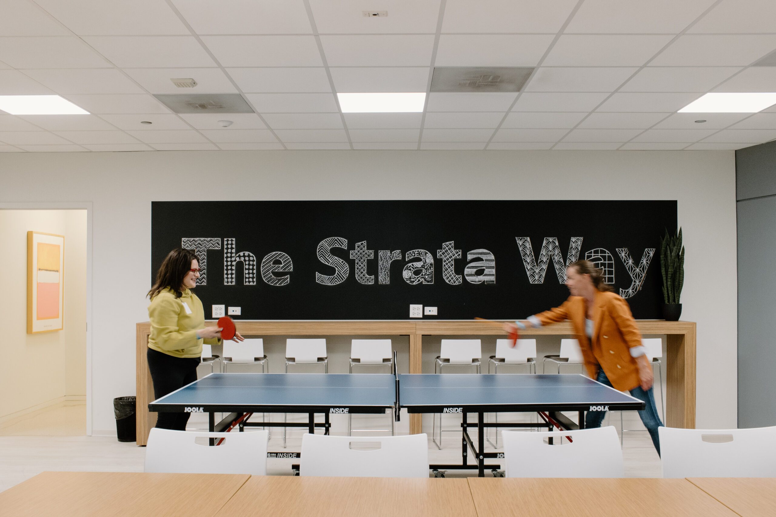 Two employees play table tennis beneath The Strata Way lettering in the Strata office