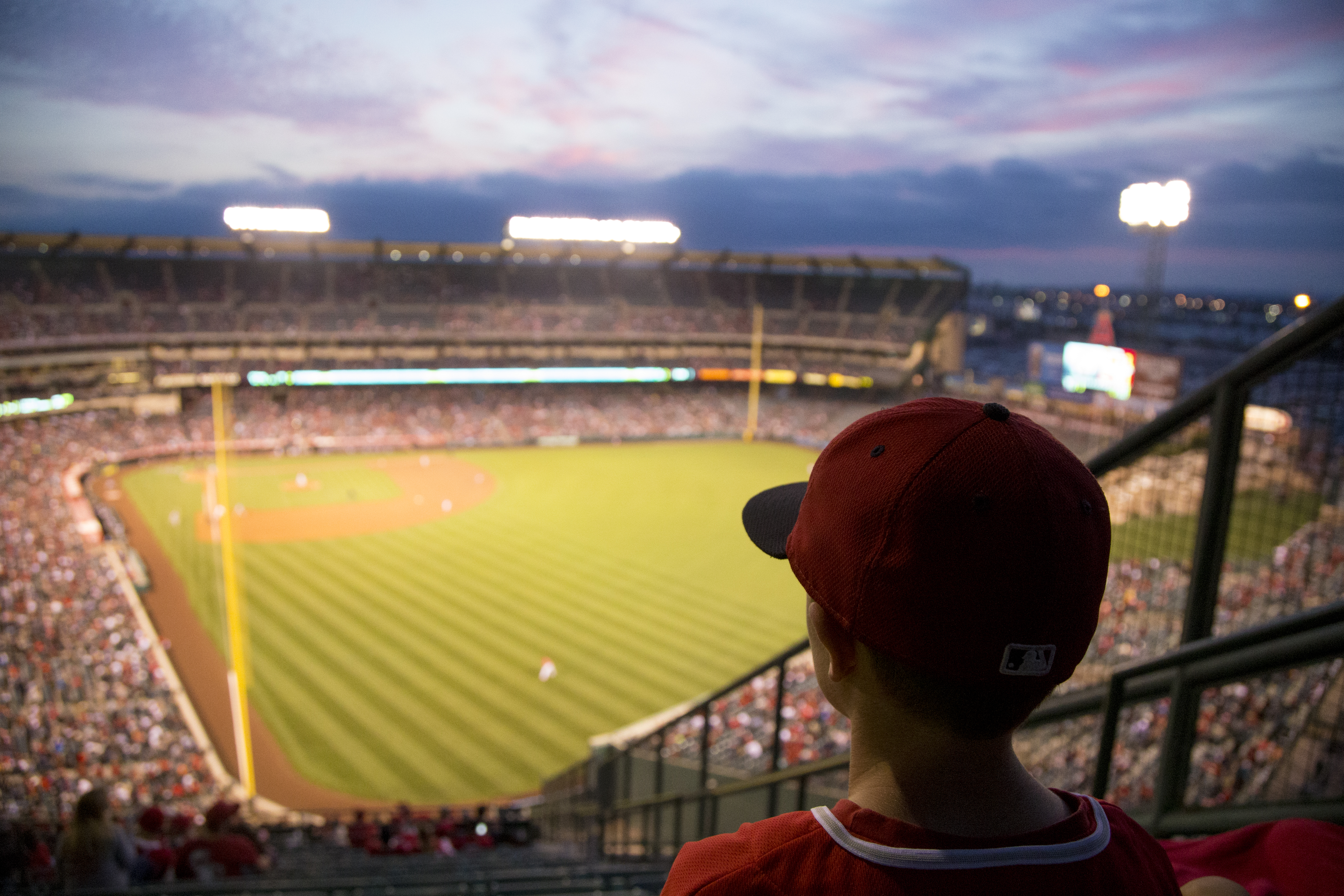 A young baseball fan in a red cap watches the field from the stands at dusk