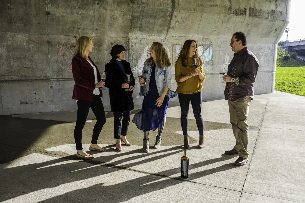 Studio 707 team holding wine glasses beneath a concrete structure, with a bottle on the pavement