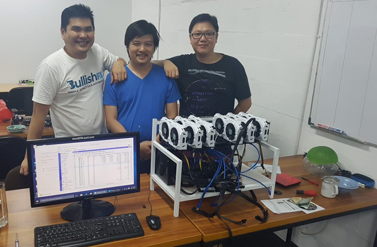 Three early Rekeningku team members standing behind a desk with a monitor and an open GPU mining rig