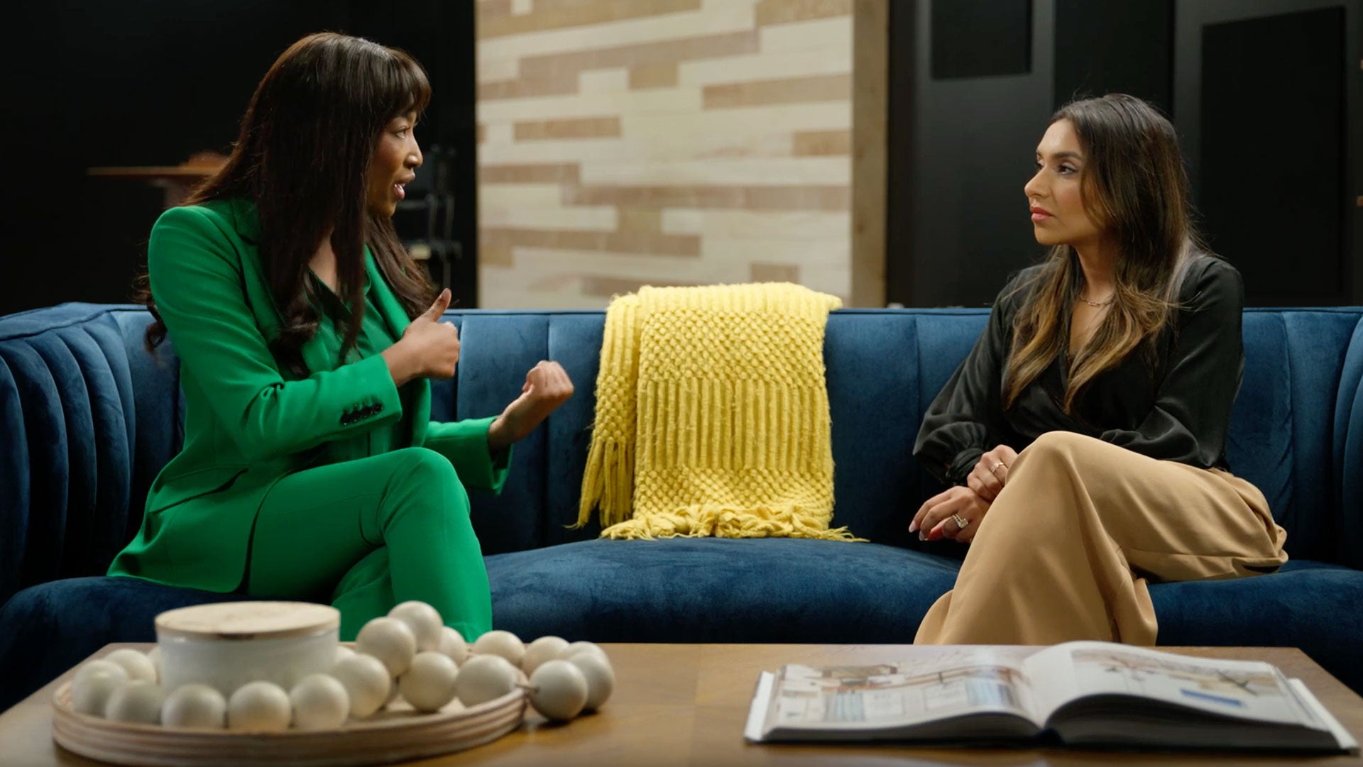 Aisha Bowe in a green suit talks with Suneera Madhani, seated at right on a blue sofa.
