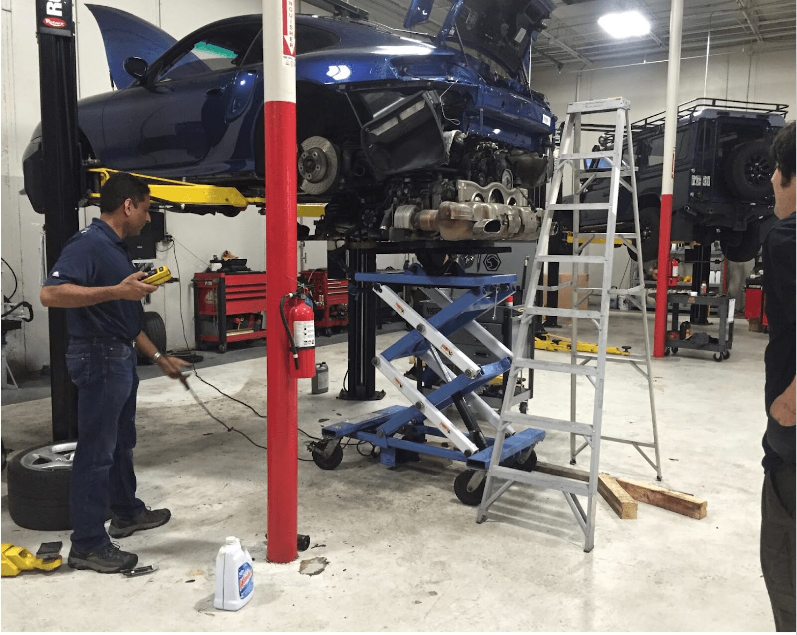 Sunil Patel at Motorwerks beside a blue car on a lift, with tools and workshop equipment around him.