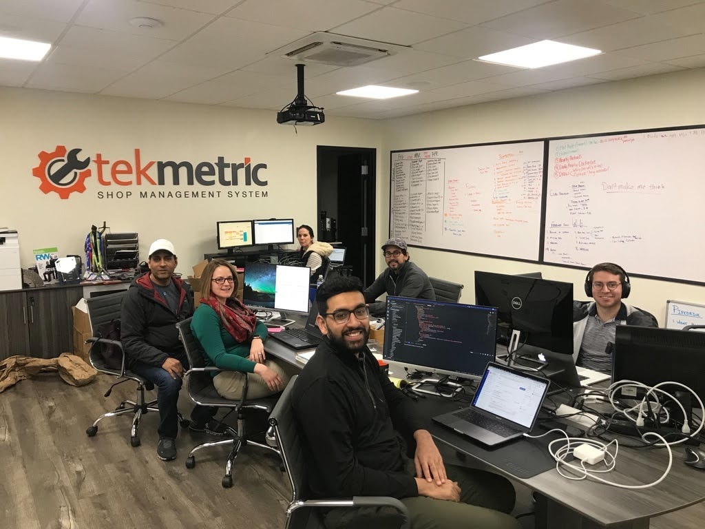 An early Tekmetric team seated together in an office filled with monitors, cables and whiteboards.