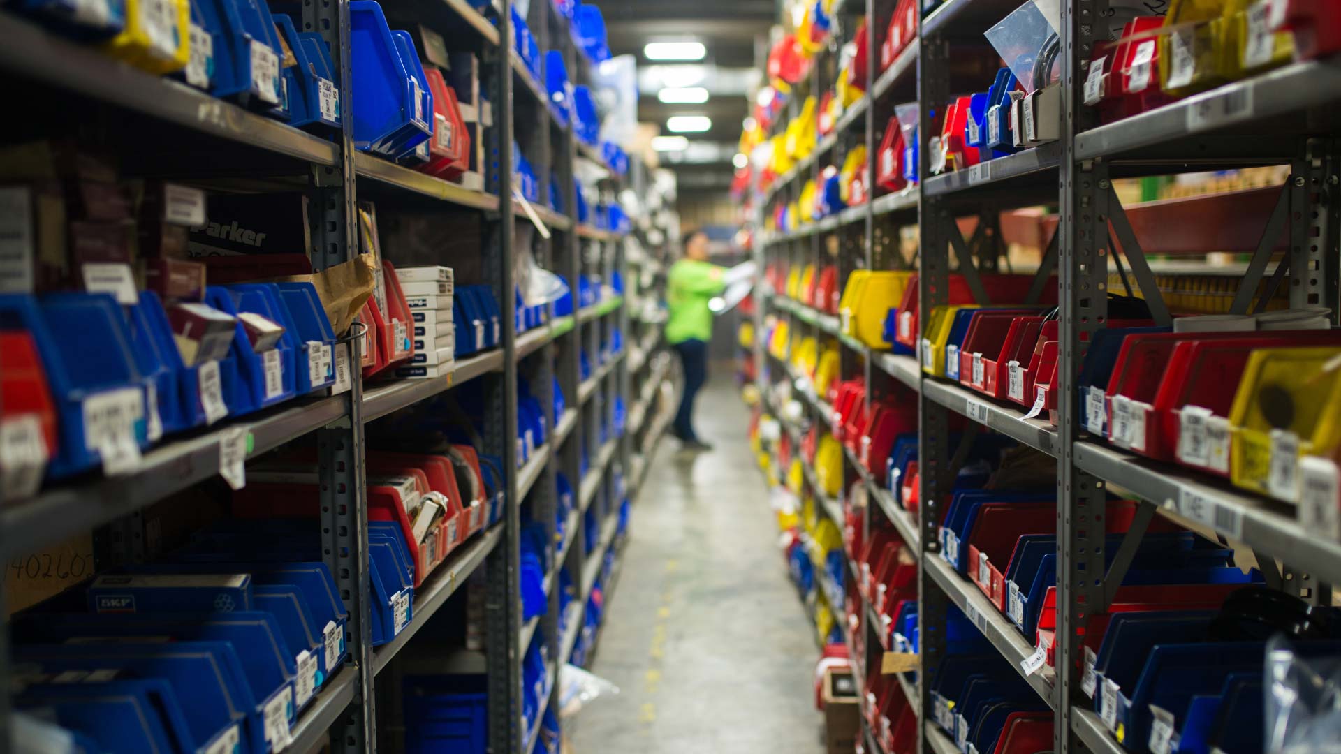 Warehouse aisle with labelled red and blue picking bins and a worker in the distance