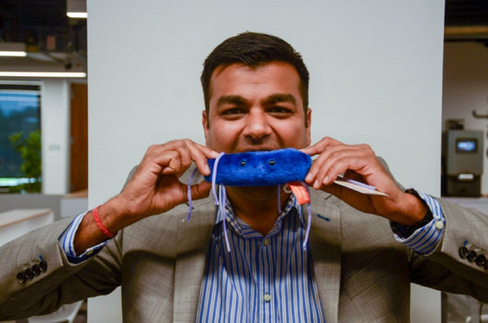 Sunny Jain in a jacket and striped shirt holding a blue plush prop in front of his face