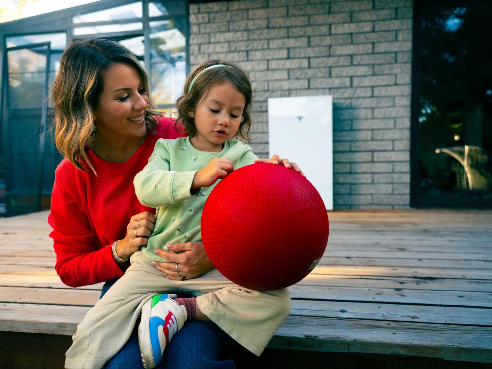 A mother and child on a deck, with a Tesla home battery mounted on the wall behind them