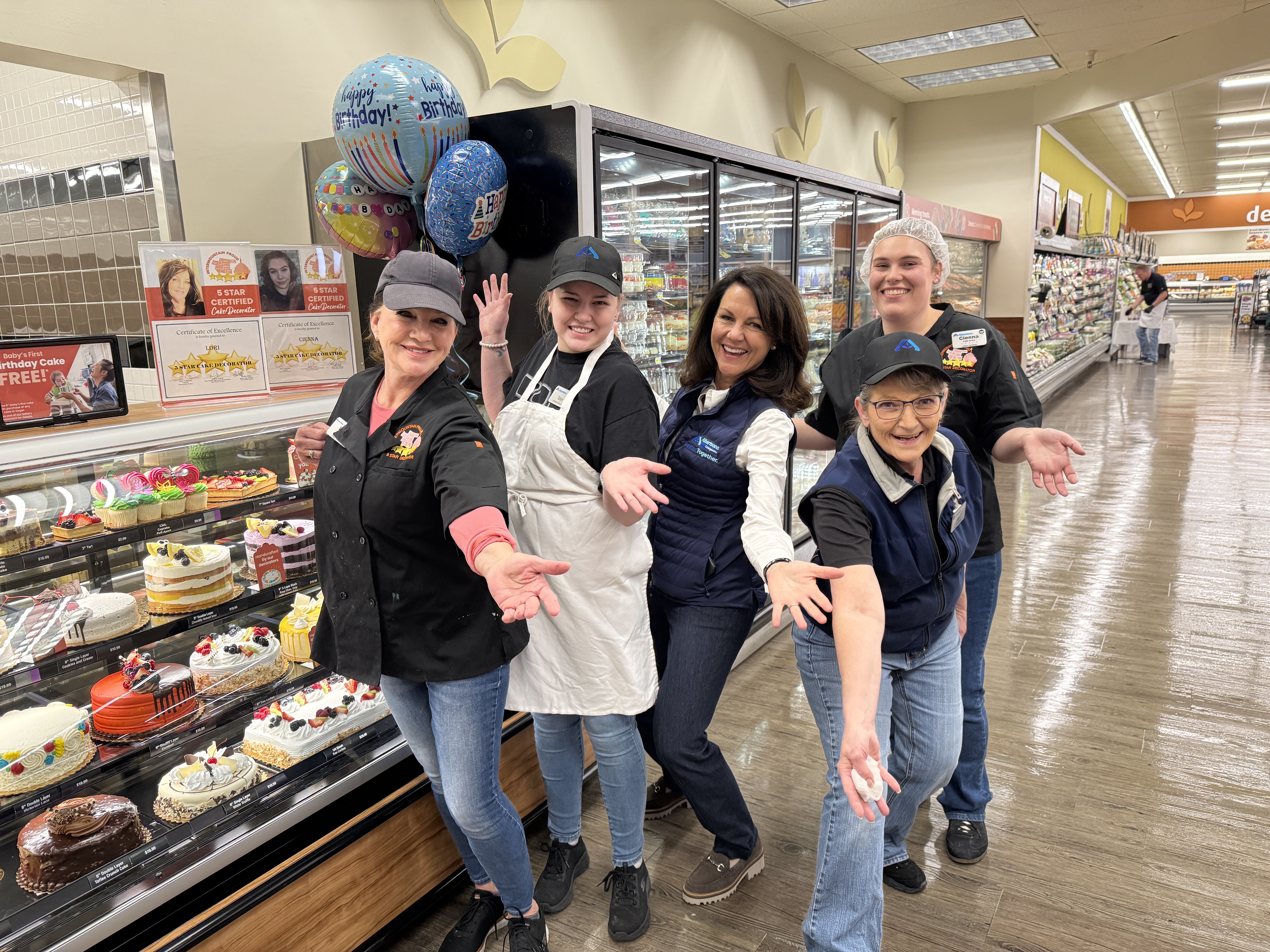 Susan Morris and four bakery associates extend their arms beside a cake display in Ammon, Idaho.