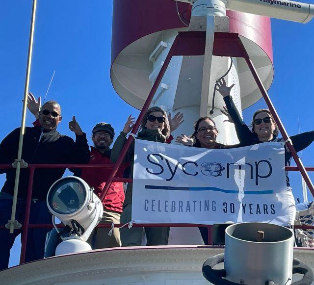 Sycomp team members aboard a boat holding a banner celebrating 30 years