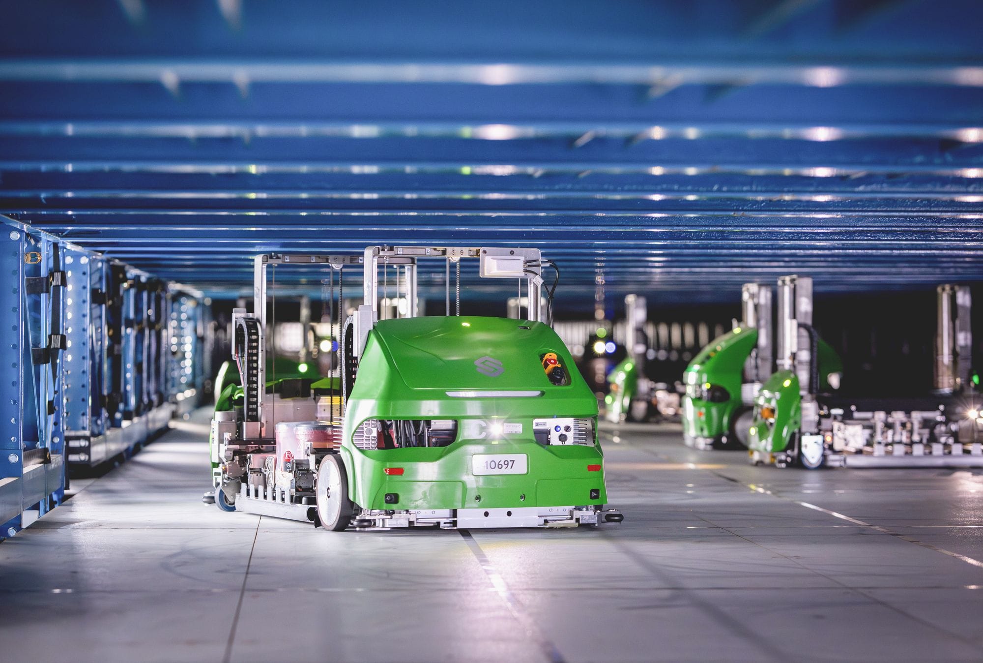 Green Symbotic autonomous robots beneath the blue steel framework of an automated storage system