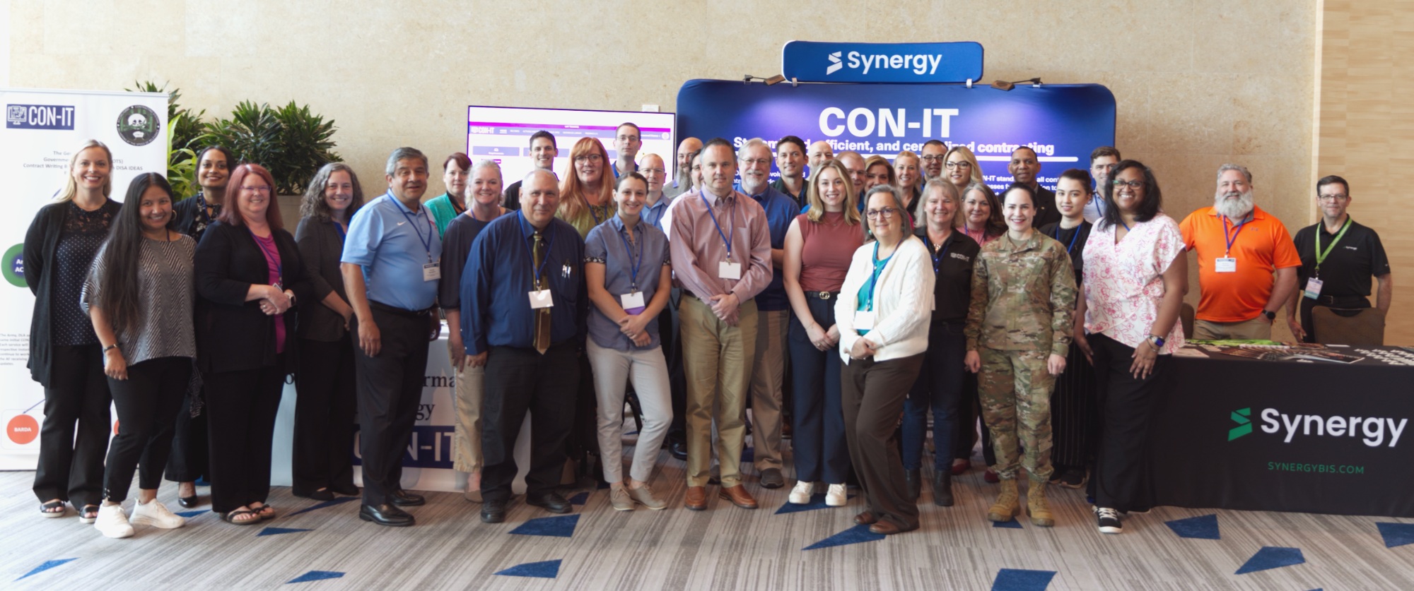 A group gathered beside Synergy and CON-IT displays at a procurement event
