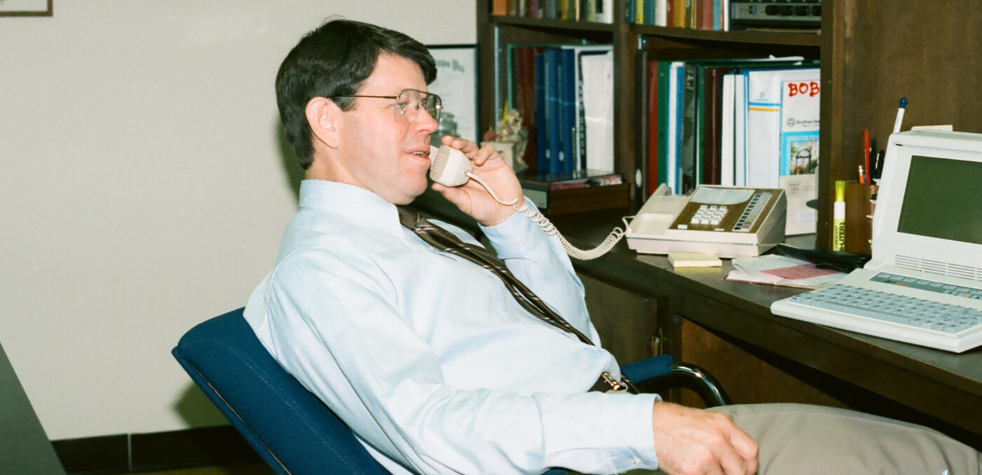 An archival SMI office photograph showing a man at a desk with a corded telephone and desktop computer