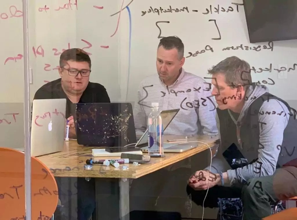Three people working around laptops behind a glass wall covered in handwritten notes