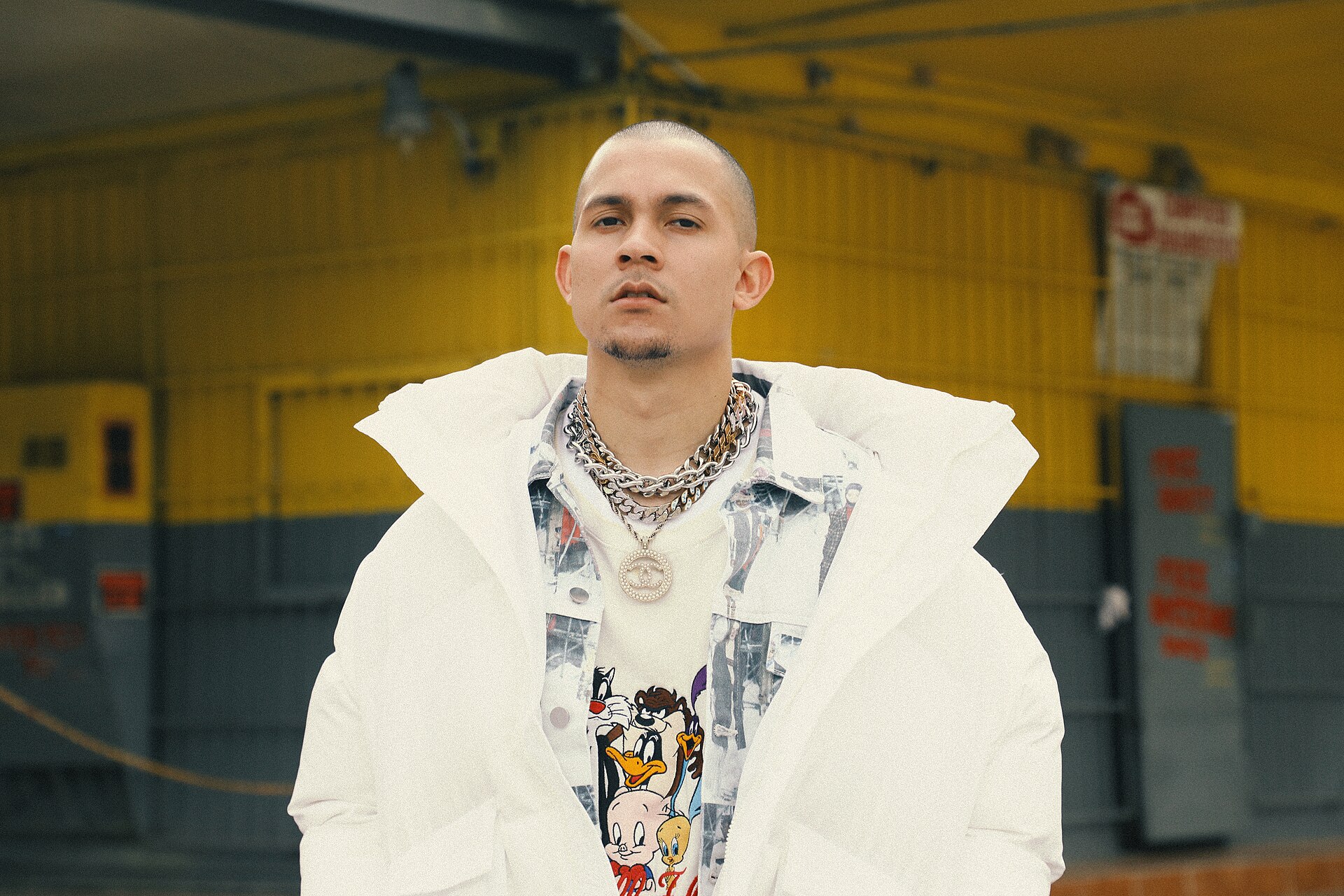Tainy wearing a white jacket in front of an industrial yellow background