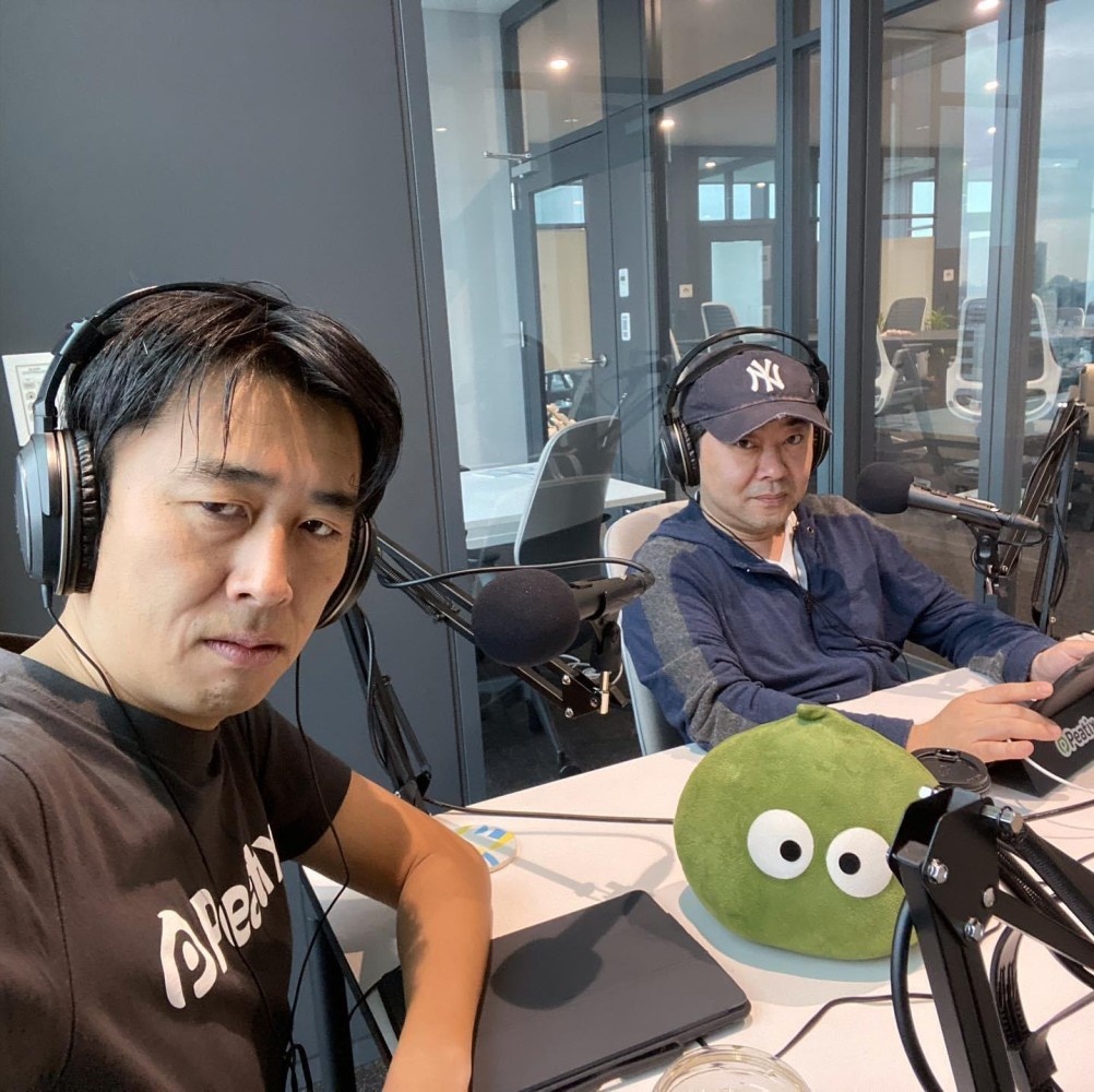 Taku Harada, at right wearing a Yankees cap, at a recording table with a colleague and Peatix’s green mascot.