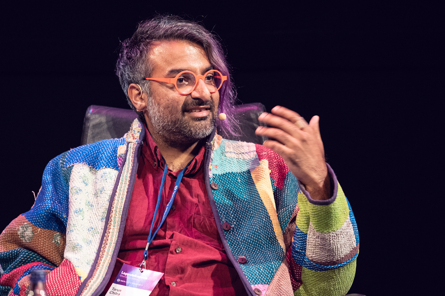Tarun Chitra speaking onstage at Legends4Legends, wearing orange glasses and a colorful patchwork jacket